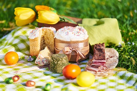 Easter cake and bright colored eggs gathered together with meat, treats and tulips on the lawn among the grassの写真素材