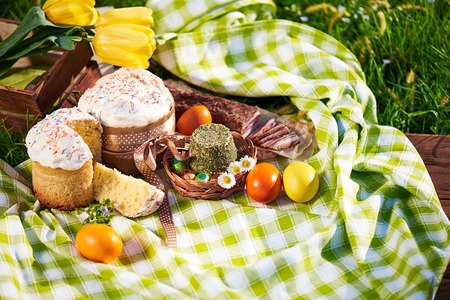 Easter cake and bright colored eggs gathered together with meat, treats and tulips on the lawn among the grassの写真素材