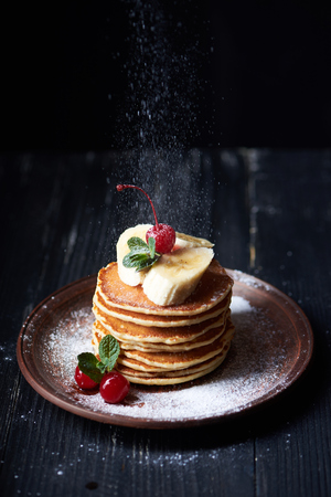 American pancakes on a plate with banana, cherry, mint and powdered sugar. Dark background. Vertical shotの写真素材