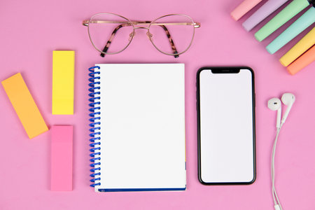 blank notebook on a pink background with pen, mobile phone with blank screen, colored post-it notes, headphones, colored markers and glassesの写真素材