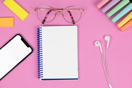 blank notebook on a pink background with pen, mobile phone with blank screen, colored post-it notes, headphones, colored markers and glassesの写真素材