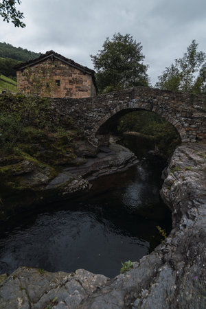 arch of a bridge over a river with dark water and a stone hut in the background with cloudy skyの写真素材