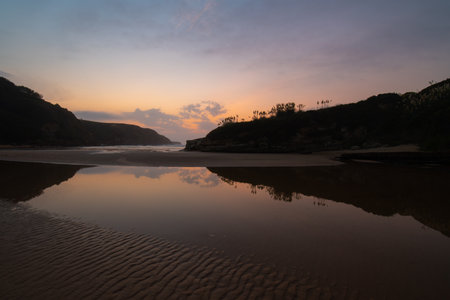 warm sunset on a beach with reflections of the sky in the water and silhouettes of vegetation on the rocksの写真素材