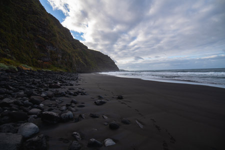 volcanic sand beach with polished rocks on a cliff with green vegetation and steps of a person who has walked on the sandの写真素材