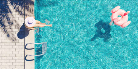 aerial view of a woman with hat sitting on the edge of the pool with palm tree shadow and flamingo...