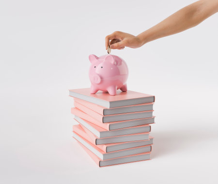 A hand placing a coin into a pink piggy bank situated on top of a stack of pink-covered books on an isolated white background. Saving and education concept. 3d rendering.の写真素材