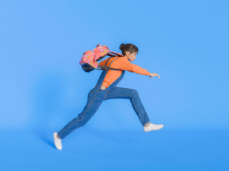 A young girl with a colorful backpack leaps energetically against an isolated blue background. Concept of freedom and adventure. 3d renderingの写真素材
