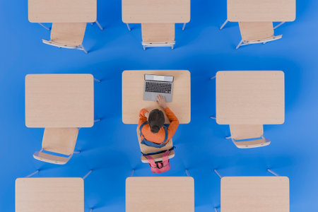 A top view of a young girl studying alone at a desk with a laptop in a classroom. Surrounded by empty desks and chairs on an isolated blue background. Concept of learning and focus. 3d renderingの写真素材