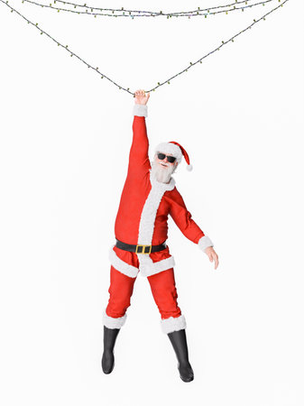 Santa claus wearing sunglasses is hanging from christmas lights with a big smile on his face. 3d renderingの写真素材