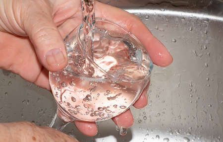 An elderly woman's hands are rinsing a glass in running clear water from the tap in the sink.の写真素材