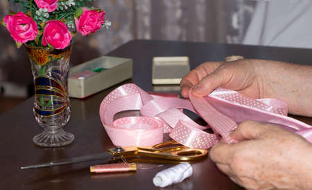 An elderly woman dressmaker, measuring out a pink ribbon.