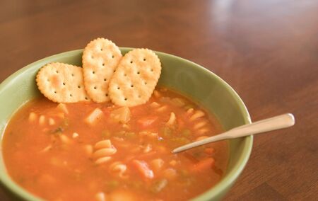 Steaming hot bowl of vegetable and pasta soup with spoon and three crackers on a wooden table.の写真素材