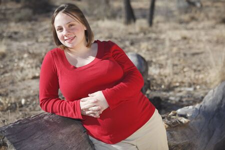Young 8 months pregnant young woman standing next to a log outdoors wearing a red shirt の写真素材