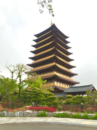 A tall wooden pagoda in Korea stands peacefully surrounded by green trees and flowers. The traditional architecture and garden setting evoke calm and cultural heritage.の写真素材