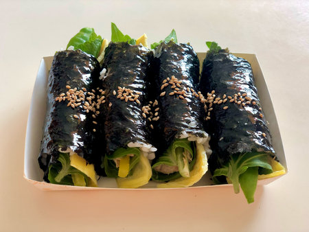 A close-up of four neatly rolled Korean-style seaweed rice rolls (gimbap or kimbap) filled with fresh vegetables and eggs, topped with sesame seeds.の写真素材