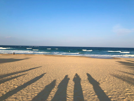 A scenic view of a peaceful sandy beach with waves gently crashing on the shore under a clear blue sky. Long shadows of people stretch across the sandの写真素材