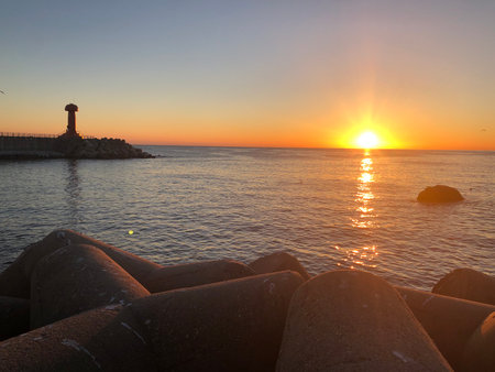The golden sun rises over a calm ocean, casting light on the breakwater and tetrapods. A tranquil seascape blending nature and coastal structures.の写真素材