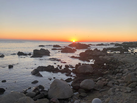 The sun rises over a rocky shoreline, casting a golden glow on the ocean and scattered boulders. A peaceful moment capturing the beauty of nature.の写真素材