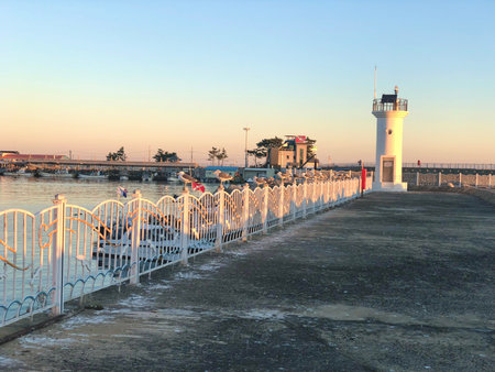A peaceful coastal pier lined with a white fence leads to a small lighthouse, gently illuminated by the setting sun.の写真素材