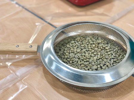 Raw green coffee beans prepared for roasting, placed in a perforated metal pan with a wooden handle. Ideal for use in coffee education, roasting guides, and artisan brewing contentの写真素材