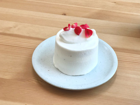 A small white frosted cake topped with red rose petals, served on a ceramic plate over a wooden table. Perfect for dessert-themed content, minimalist bakery branding.の写真素材