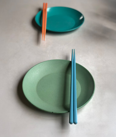 A modern minimalist table setting featuring two plates in pastel green and teal, each paired with vibrant chopsticks in blue and orange.の写真素材