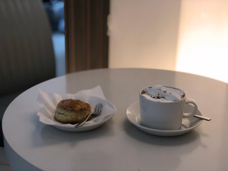 A cup of coffee topped with whipped cream, served with a freshly baked scone on a white round table in a cozy cafe.の写真素材