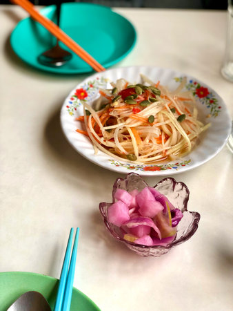 A vibrant Asian-style table setting featuring a plate of fresh papaya salad garnished with pumpkin seeds, served alongside a small bowl of pink pickled radish.の写真素材