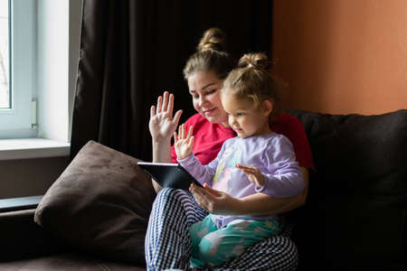 Mother and doughter chating, make video call, on tablet computer at home. Little girl waves her hand...