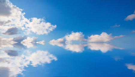 A clear blue sky with fluffy white clouds reflects in calm water below, creating a serene and picturesque scene. The sky and clouds are mirrored perfectly in the water, creating a peaceful and tranquil atmosphere.の素材