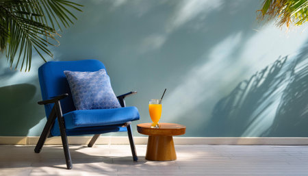 A blue armchair sits in a bright room with a large palm plant. A straw hat and an orange throw blanket rest on the chair. A side table with a glass of water and a bottle of orange juice sits nearby.の素材