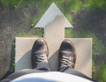 A person is positioned at the beginning of a smooth, curving pathway that leads up to a large, upward-pointing arrow. The pathway and arrow are rendered in a minimalistic blue tone, creating a serene yet motivational atmosphere. The individual appears contemplative, gazing thoughtfully at the arrow that symbolizes direction and progress, suggesting a journey towards success.の素材