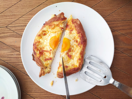 A freshly baked bread filled with cheese and topped with two sunny-side-up eggs is served on a white plate. A knife and spatula are nearby for easy serving.の写真素材