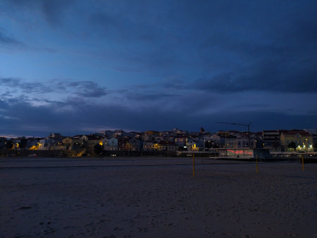 Serene twilight scene on a sandy beach, overlooking a quaint coastal town bathed in the soft glow of evening lights.の写真素材