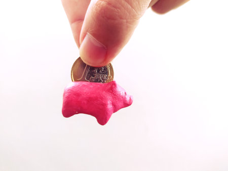 Hand putting a coin into a tiny pink piggy bank. Concept of savings, investment, finance, and economic security. Metaphor for childhood, planning, and future.の写真素材