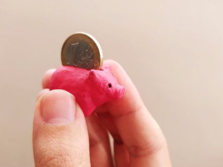 Hand holding a tiny, pink piggy bank with a coin being inserted. Illustrates savings, investment,...
