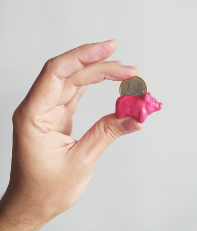 Hand holding a tiny pink piggy bank with a coin. Symbolizes savings, finance, investment, and...