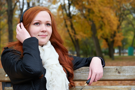 redhead girl listen music on headphones in city park, fall seasonの写真素材