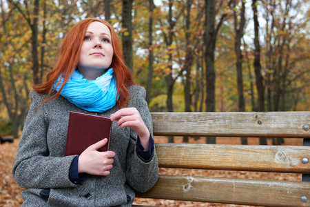 redhead girl with book sit on a bench in city park, fall seasonの写真素材