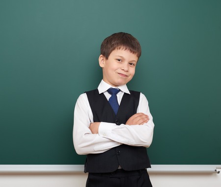 school student boy posing at the clean blackboard, grimacing and emotions, dressed in a black suit,...