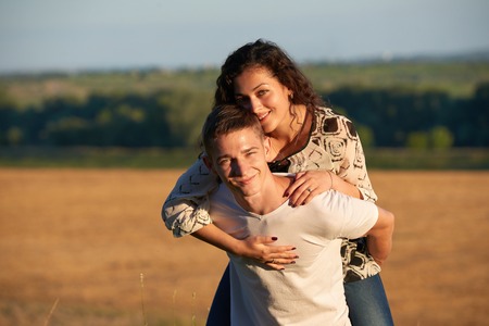 holidays, hiking, love and friendship concept - smiling couple having fun over evening sky background, summer season, girl riding on man backの写真素材