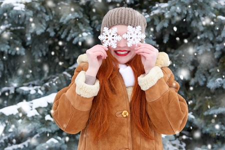 beautiful woman on winter outdoor posing with big snowflake toys, holiday concept, snowy fir trees in forest, long red hair, wearing a sheepskin coatの写真素材