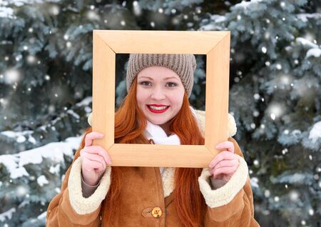 beautiful woman portrait on winter outdoor, look through wooden frame, snowy fir trees in forest, long red hair, wearing a sheepskin coatの写真素材