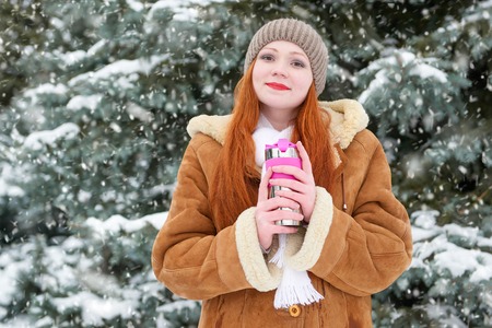 beautiful woman drinking a hot drink and keep warm on winter outdoor, snowy fir trees in forest, long red hair, wearing a sheepskin coatの写真素材