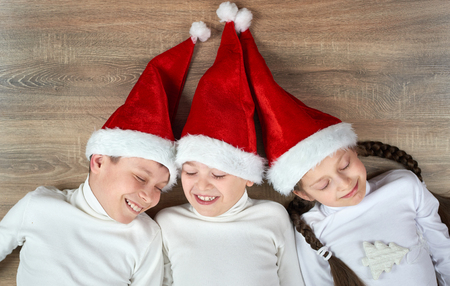 three kids in Santa hats lying on wooden background, having fun and happy emotions, winter holiday conceptの写真素材