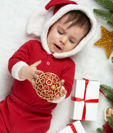 baby portrait in christmas decoration, dressed as Santa, lie on fur near fir tree and play with gifts, winter holiday conceptの写真素材