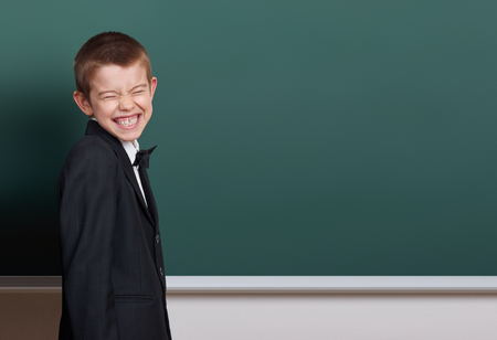 elementary school boy make faces near blank chalkboard background, dressed in classic black suit, one pupil, education conceptの写真素材