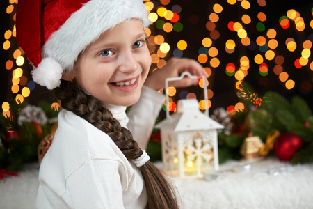 child girl portrait with christmas decoration, dark background with lights, face expression and happy emotions, dressed in santa hat, winter holiday conceptの写真素材