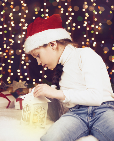 girl child posing with christmas decoration on dark background, illuminated lights and bokeh, face closeup, dressed in santa hat, winter holiday concept, xmas treeの写真素材