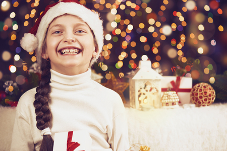 girl child posing with christmas decoration on dark background, illuminated lights and bokeh, face closeup, dressed in santa hat, winter holiday concept, xmas treeの写真素材
